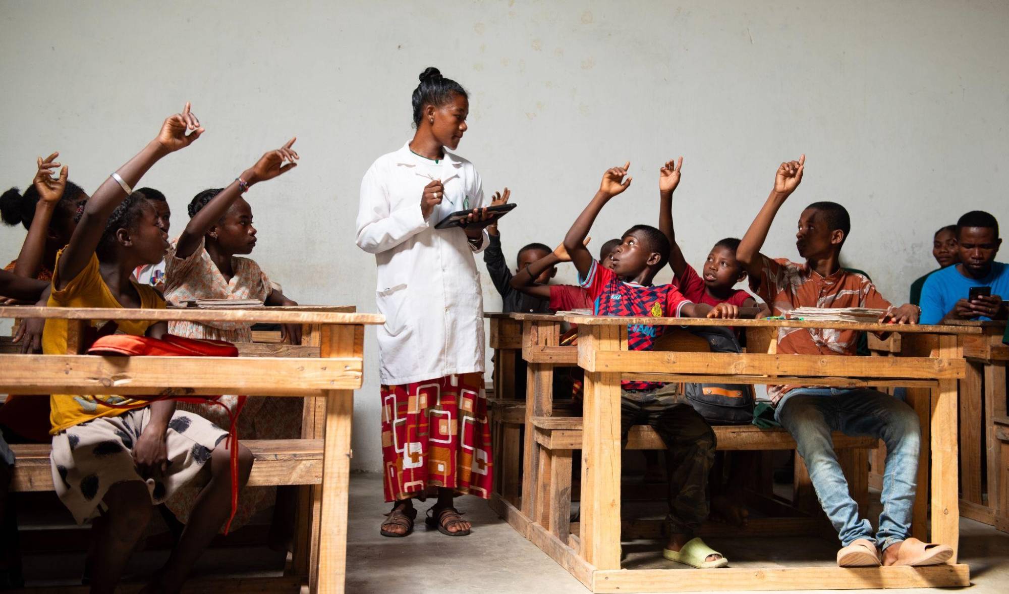A teacher holding a tablet and students raising their hands